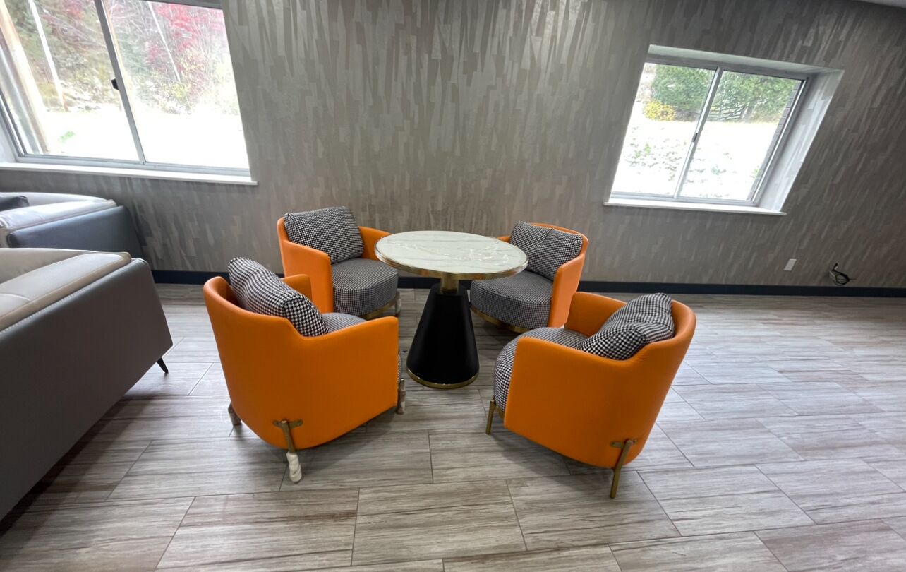 A modern living room with a grey sofa, two orange armchairs, a round coffee table, and a view of trees through large windows.