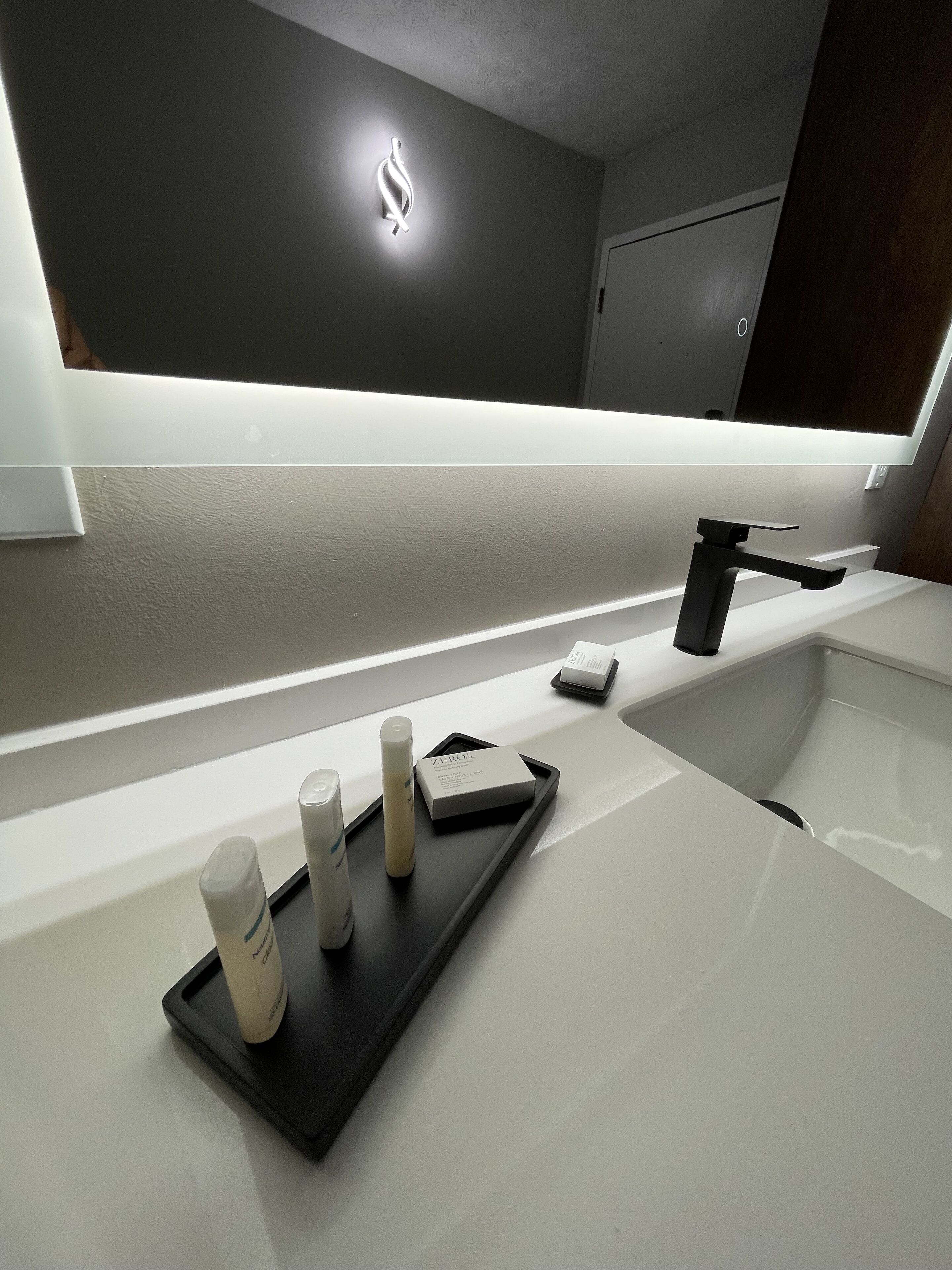 A modern bathroom with a sleek white sink, a black tray with toiletries, and a wall-mounted light fixture.