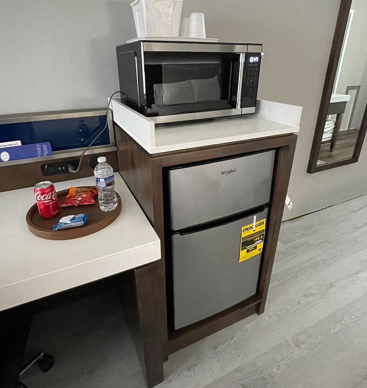 Una kitchenette con un microondas sobre un refrigerador, una mesita con una botella de agua, una lata de Coca-Cola y una bolsita de snack.