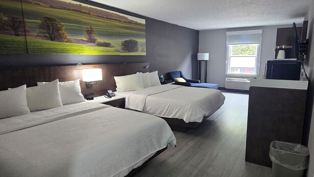 A hotel room with two beds, a desk, a chair, a lamp, and a painting of a landscape.