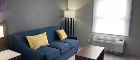 A modern living room with a blue sofa, two yellow cushions, a floor lamp, a wooden coffee table, and a large window with blinds.