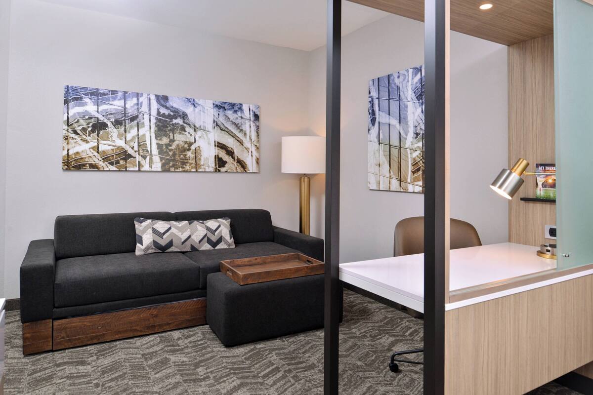 A modern hotel room with a sofa, a wooden side table, a desk with a lamp, and two wall-mounted artworks.