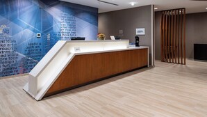 Un comptoir d’accueil moderne, doté d’un revêtement en bois et d’un plan de travail blanc, situé dans un hall d’entrée avec un mur aux motifs géométriques et une cloison en bois.