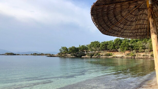 A beach with clear water, a thatched umbrella, and a rocky shoreline with trees. - Family friendly house close to the beach (Κριθάρια)