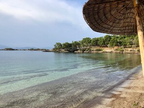 Una playa con aguas cristalinas, un paraguas de paja y una costa rocosa con árboles.