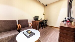 Ein kompaktes Hotelzimmer mit einer Sitzecke, einem kleinen runden Tisch, einem Schreibtisch mit Computer und einer Küchenzeile.