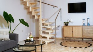 Un salon moderne avec un escalier en colimaçon, un téléviseur à écran plat, un canapé gris, une table basse ronde en verre et un tapis en rotin.