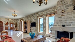 A spacious living room with stone walls, a fireplace, and large windows offering a mountain view. - HECTORAS VILLA in Plaka-BY APOKORONAS VILLAS (Πλάκα)