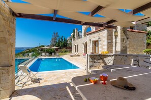 A poolside area with a stone pool, lounge chairs, and a table with fruits and a hat. - HECTORAS VILLA in Plaka-BY APOKORONAS VILLAS (Πλάκα)