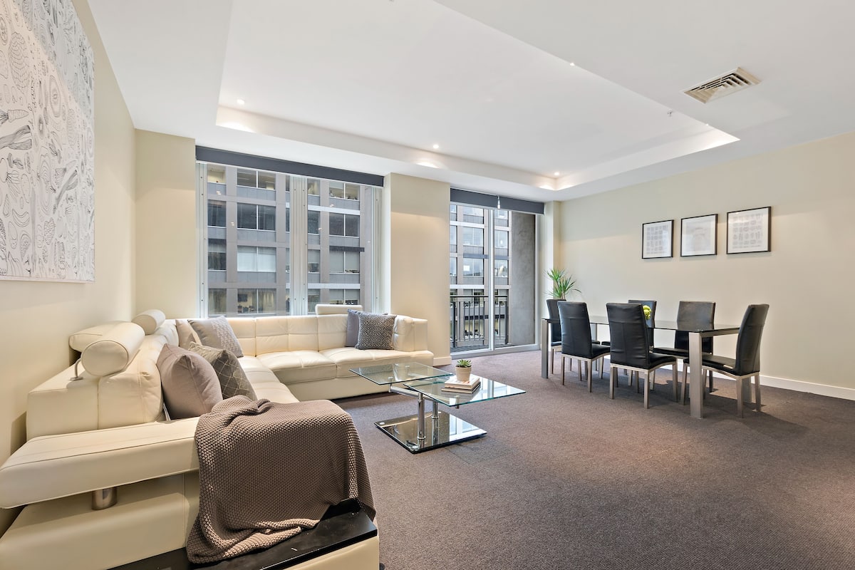 A modern living room with a white sofa, glass coffee table, and a dining area with a table and chairs.