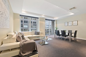 A modern living room with a white sofa, glass coffee table, and a dining area with a table and chairs. - Nook Melbourne - Collins Street (Melbourne)