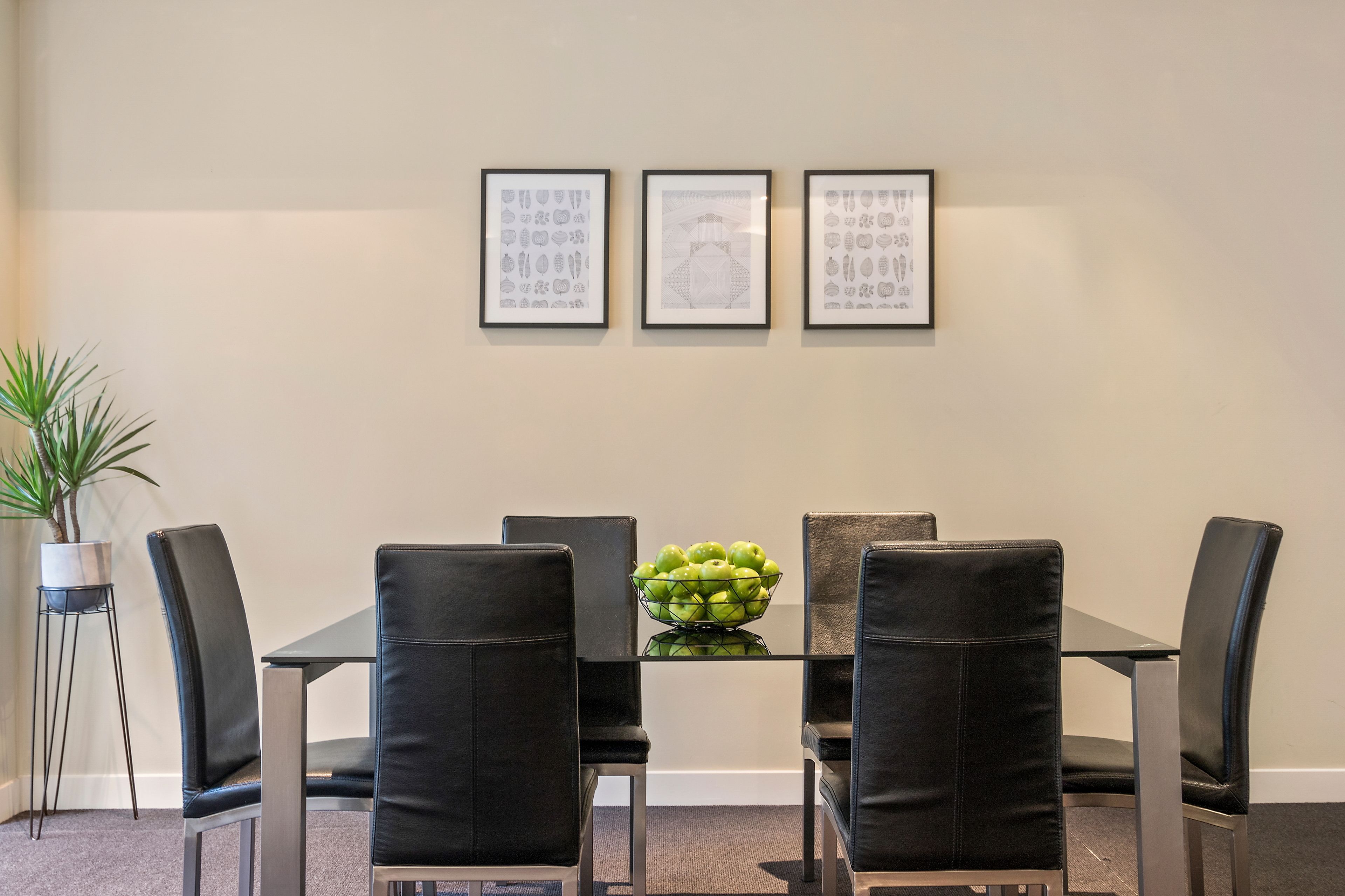 Un coin repas avec une table en verre, des chaises noires et un saladier de pommes vertes. Trois tableaux encadrés sont accrochés au mur.