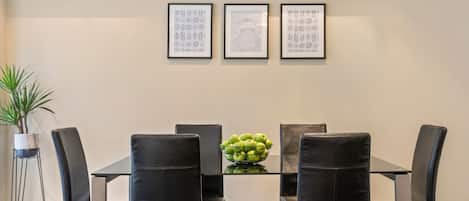 Un coin repas avec une table en verre, des chaises noires et un saladier de pommes vertes. Trois tableaux encadrés sont accrochés au mur.