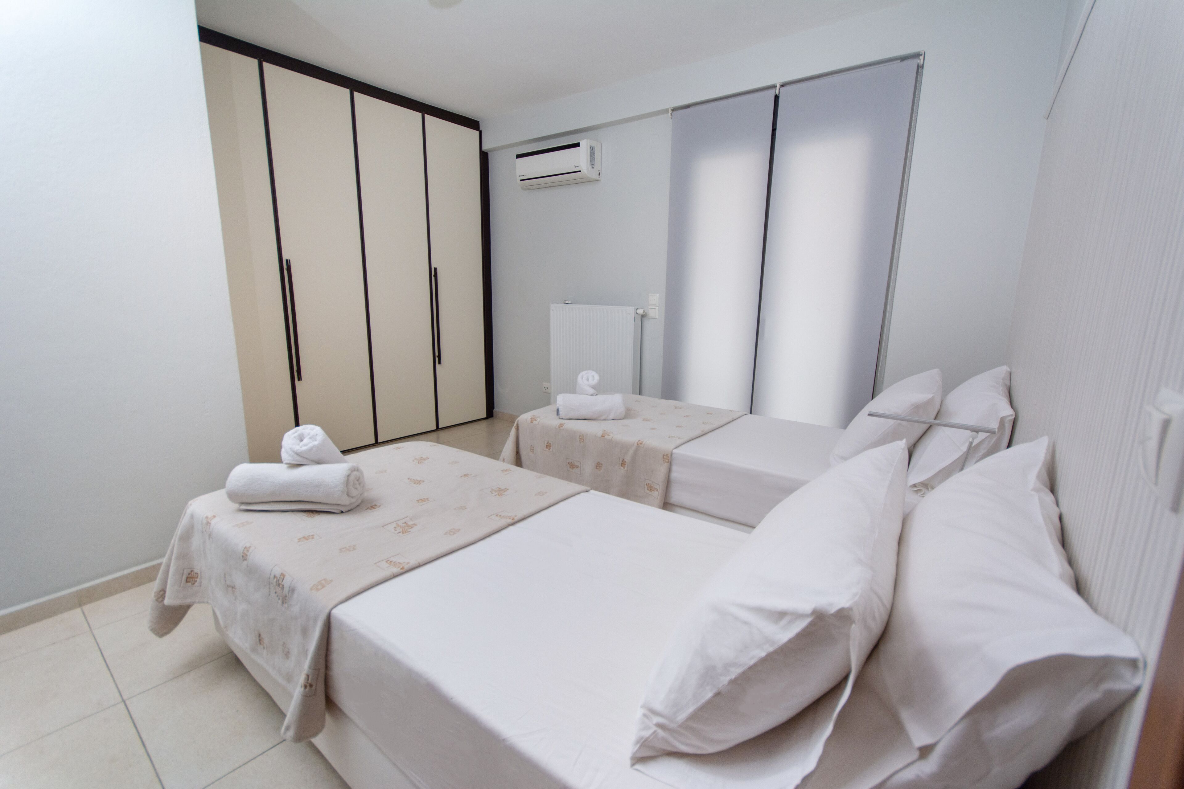 A hotel room with two beds, a wardrobe, an air conditioning unit, and a radiator.