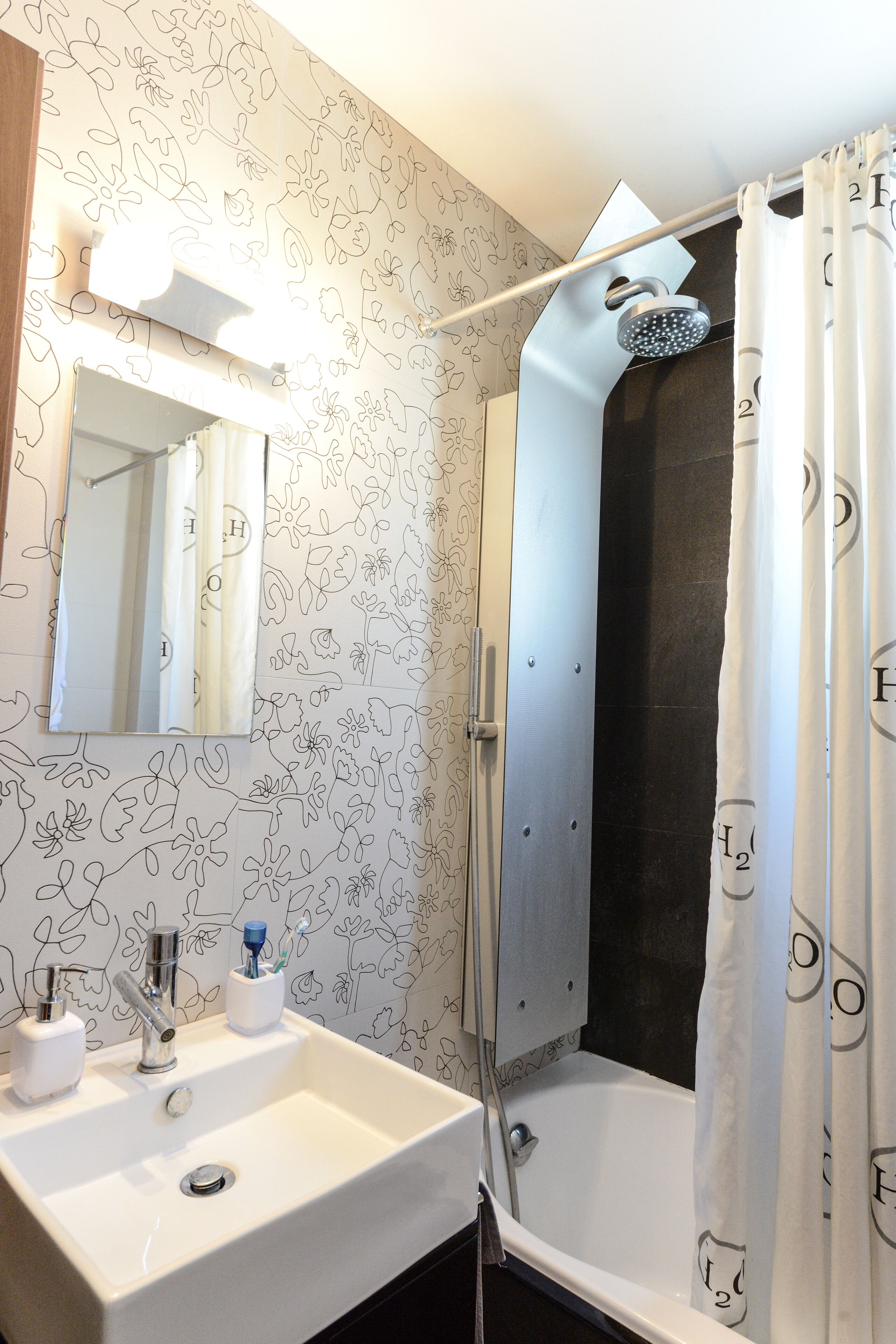 Un baño con un lavabo blanco, un espejo, una ducha con puerta de cristal y una cortina de ducha estampada.
