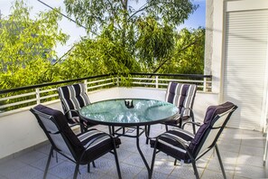 A round glass table with four chairs, a white railing, and a view of trees and a building in the background.