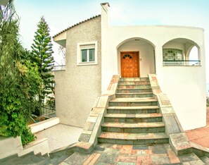 A building with a staircase leading to a wooden door, surrounded by greenery.