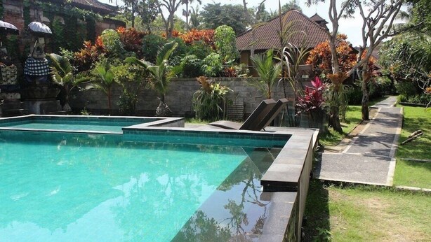 A rectangular swimming pool with clear water, surrounded by a stone wall and a grassy area with various plants and trees. - agung trisna bungalows (Ubud)