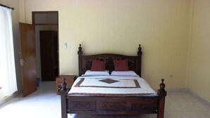A bedroom with a wooden bed, a nightstand, a door, and a light switch on the wall. - agung trisna bungalows (Ubud)