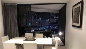 A modern dining area with a large table and chairs, a decorative bowl, and a view of a cityscape at night. - Large Apartment at Oracle Resort (Broadbeach)