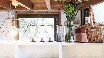 Una cocina con vigas de madera, lámparas colgantes, un ventanal con vistas, un florero con flores y estantes con frascos y vasos.