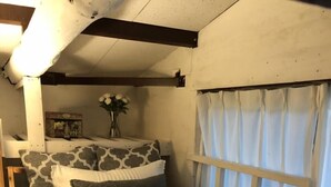 Une chambre à coucher avec un lit, des oreillers, un coussin décoratif noué, une table de chevet, un vase avec des fleurs et une fenêtre avec des rideaux.