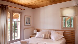 A bedroom with a wooden ceiling, a bed with white bedding, a painting on the wall, and a window with a view of greenery. - Summer Breeze House * with private pool * private parking * (Χανιά)