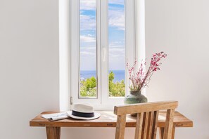 A wooden table with a newspaper, a hat, and a vase with flowers by a window with a view of greenery and the sea.