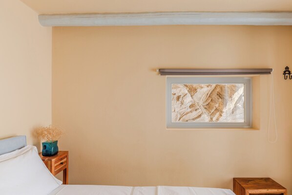 A bedroom with a bed, a nightstand with a vase, a small wooden table, a window with a view, and a wall-mounted light fixture.