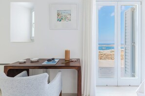 A room with a wooden table, a chair, a candle, and a framed painting. A door leads to a balcony with a view of the sea and a stone wall.