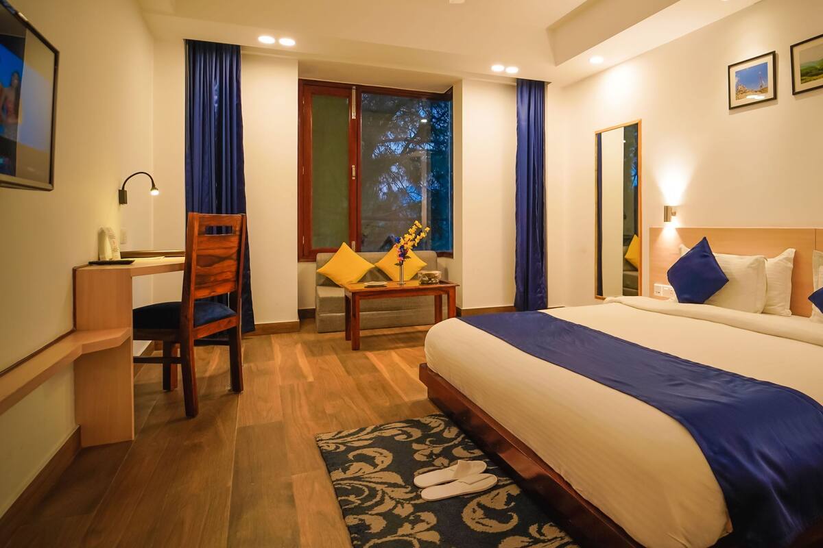 A hotel room with a bed, a desk, a chair, a small table with a vase, and a window with curtains.