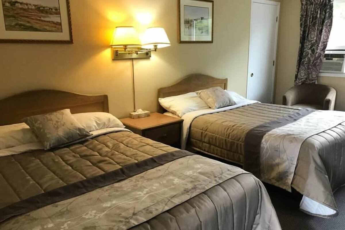 Ein Hotelzimmer mit zwei Betten, einem Nachttisch mit Lampe, einem Sessel und einem Fenster mit Vorhängen.