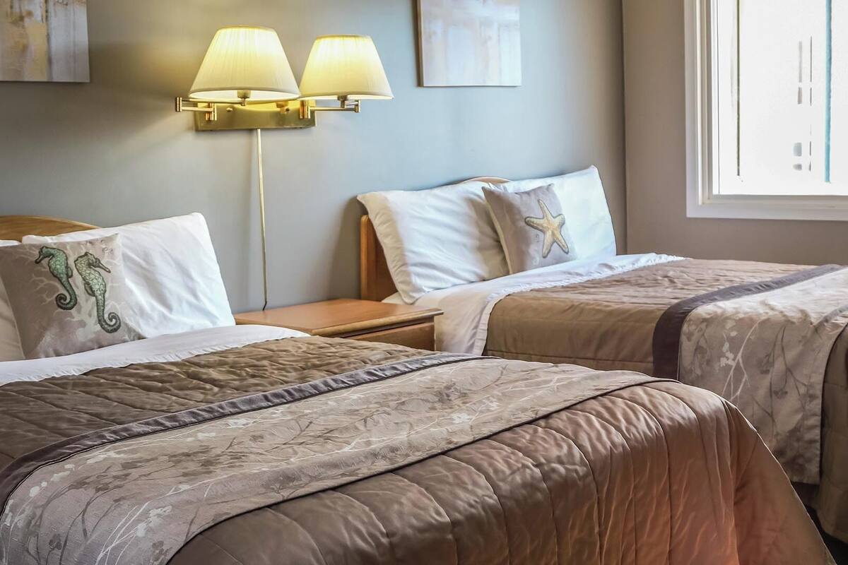 Ein Hotelzimmer mit zwei Betten, beide mit beiger Steppdecke und passenden Kissen. Dazwischen steht ein Nachttisch mit einer Lampe.