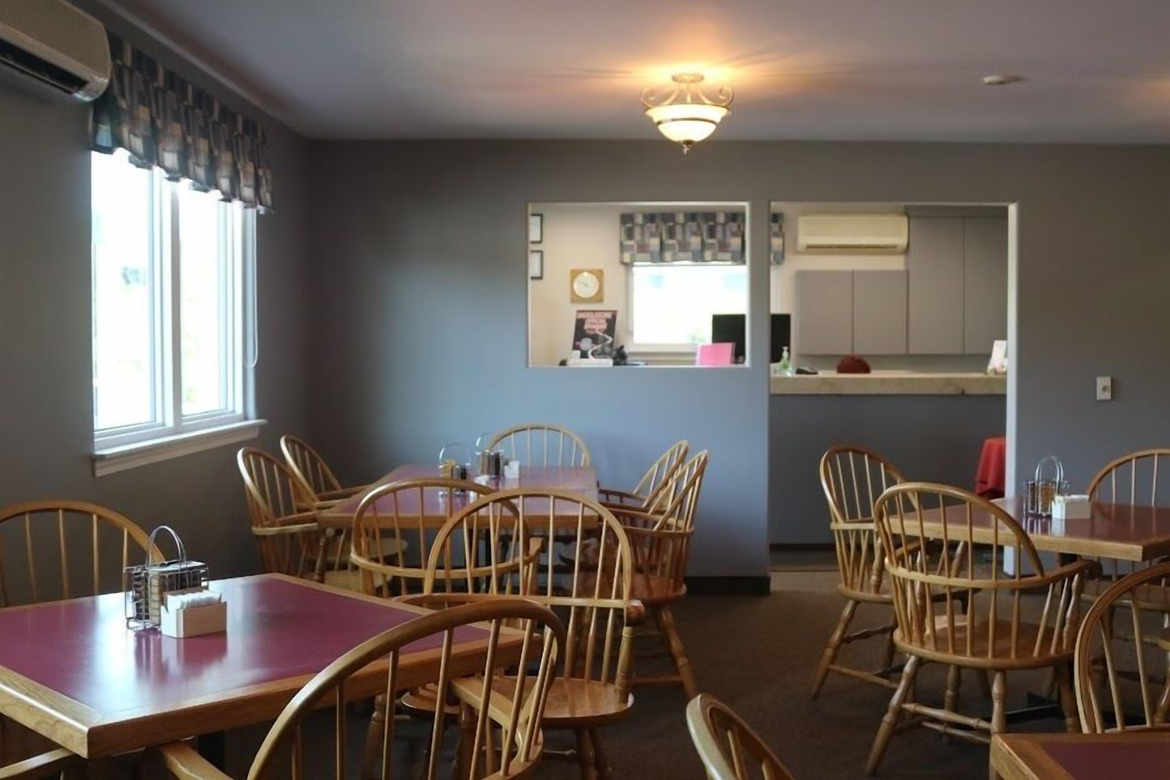 A dining area with wooden tables and chairs, a window with curtains, and a kitchen area in the background.