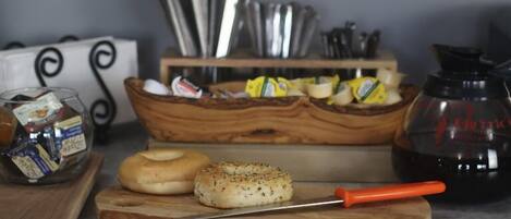 A kitchen counter with a wooden cutting board, a knife, a bagel, a jar of jam, a coffee maker, and a spice rack.