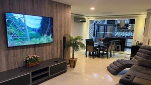 A modern living room with a flat-screen TV, a dark sofa, a dining area, and a kitchen in the background.