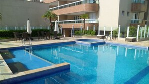 A swimming pool with a clear blue water, surrounded by lounge chairs and umbrellas, and adjacent to a multi-story building with balconies.