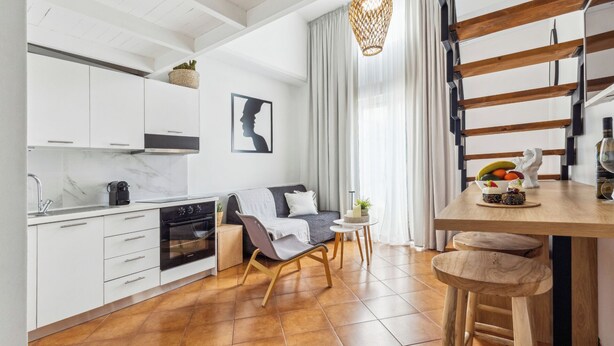 A modern kitchen with white cabinets, a built-in oven, and a coffee machine. There is a dining area with a wooden table, chairs, and a fruit bowl. A sofa and a small side table are also visible. - GGK Apartments -O- (Heraklion)