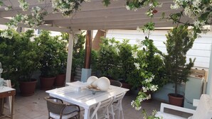 An outdoor patio with a white table and chairs, surrounded by potted plants and a pergola with white flowers. - GGK Apartments -O- (Heraklion)