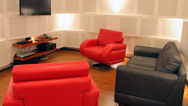 Un salon moderne avec un téléviseur à écran plat fixé au mur, un canapé rouge et un canapé gris, ainsi qu’un meuble de rangement en bois contenant des appareils électroniques.