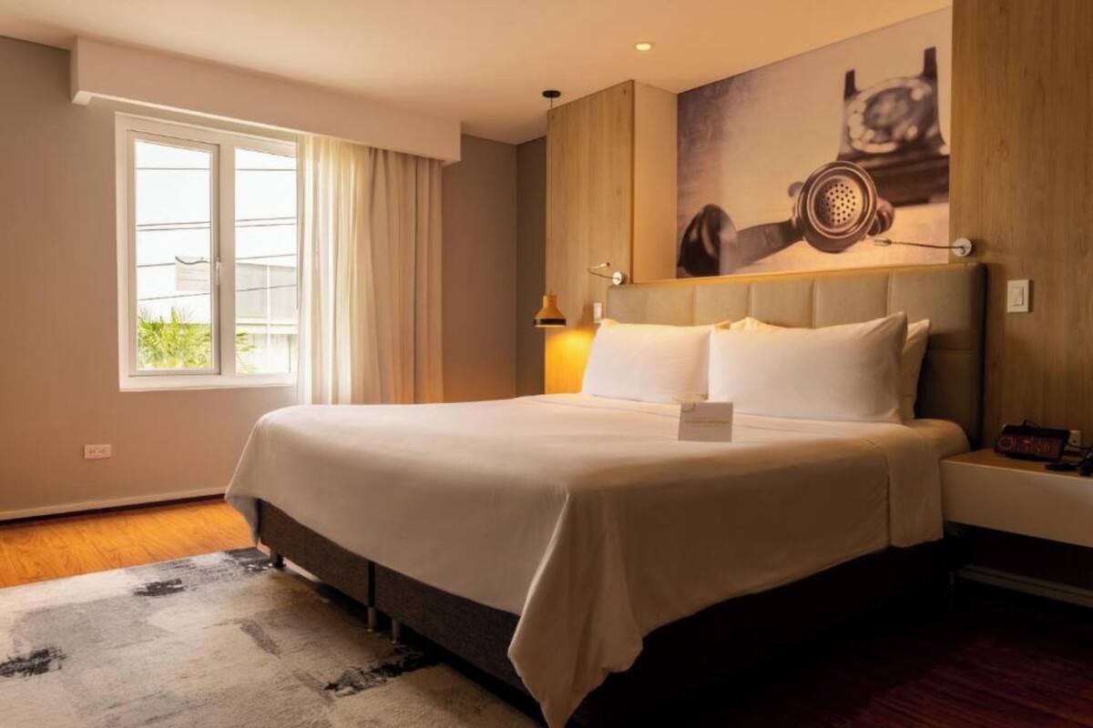 Une chambre d’hôtel avec un grand lit, une fenêtre avec des rideaux, une œuvre d’art accrochée au mur et une table de chevet avec une lampe.