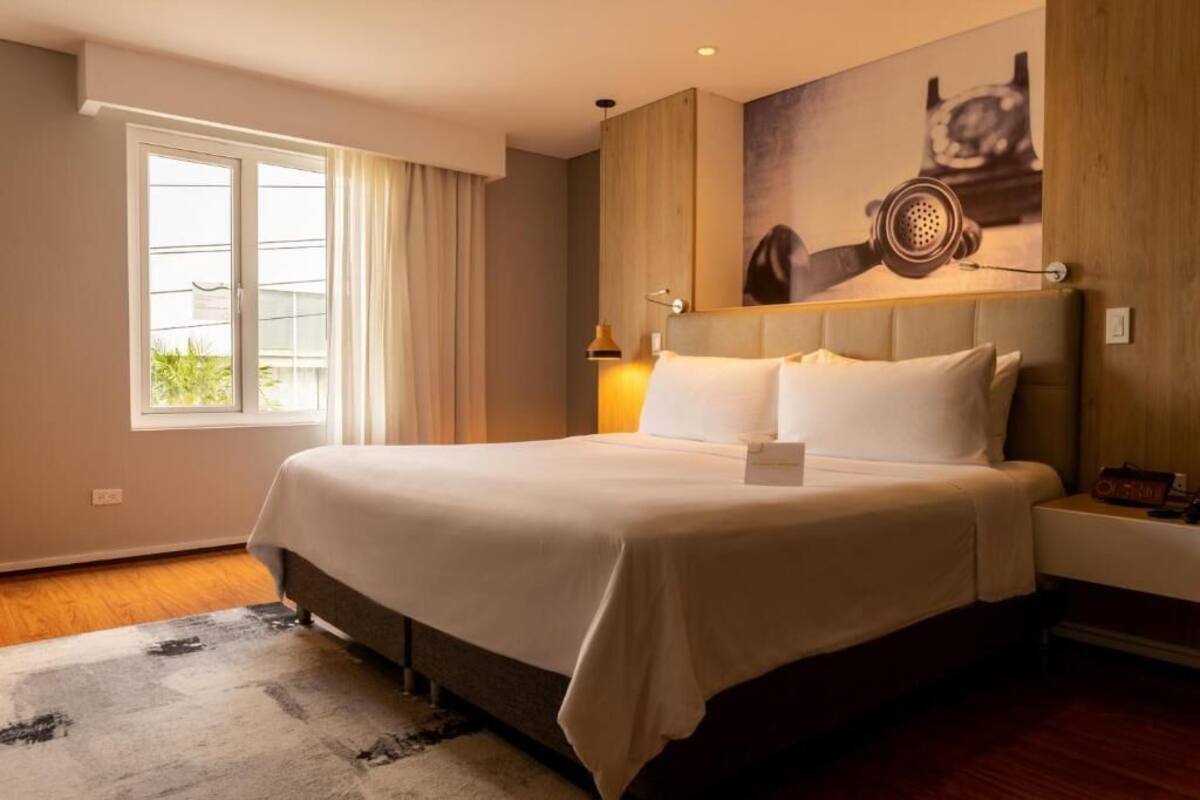 Une chambre d’hôtel moderne avec un grand lit, une fenêtre avec des rideaux, une tête de lit en bois et un tableau au mur.