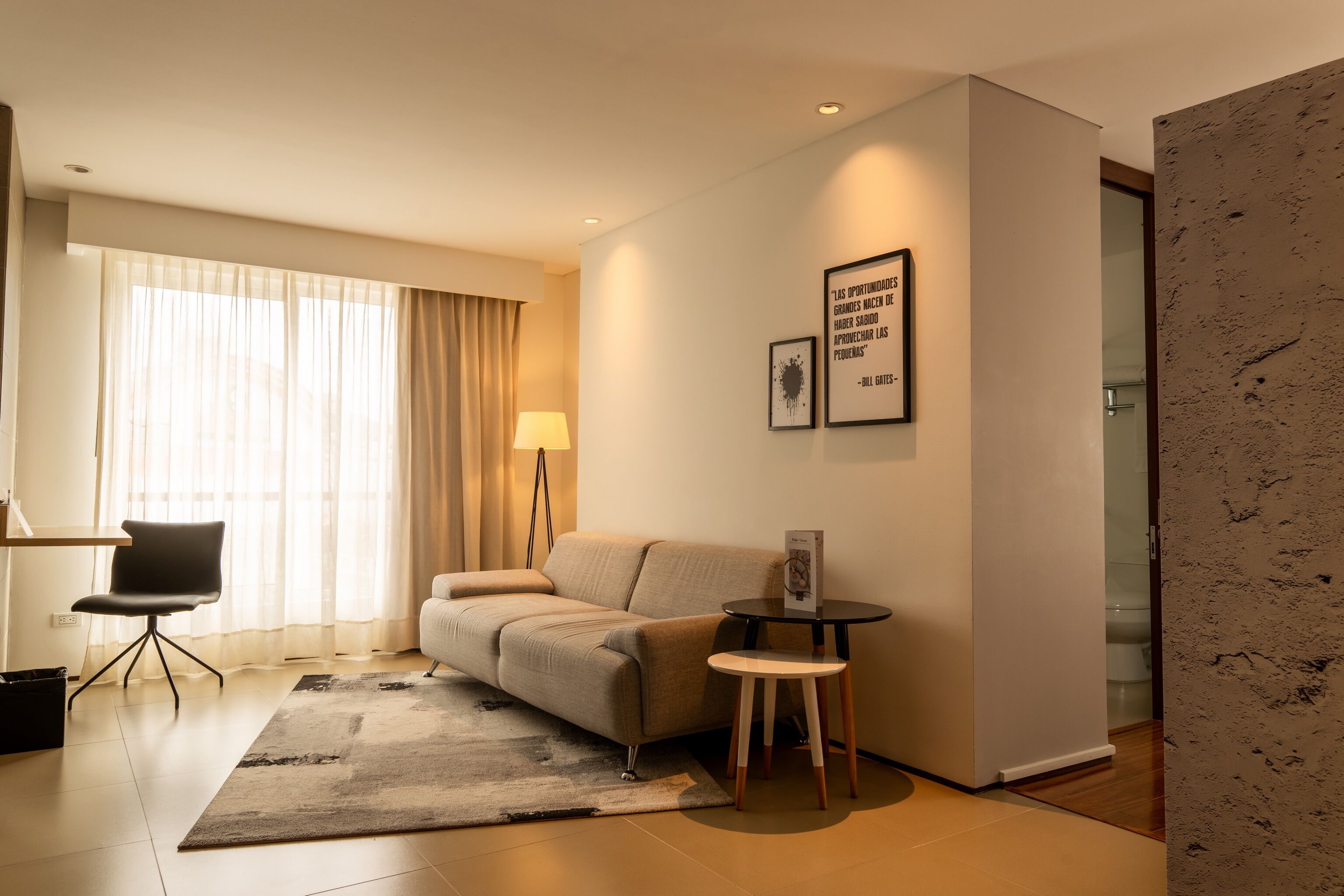 A modern hotel room with a sofa, a small table, a lamp, and a large window with curtains.