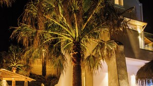 Zona de piscina con una palmera, un edificio blanco con balcón y una estructura de techo de paja.