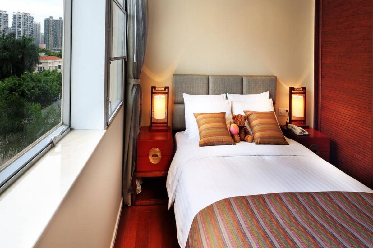 Una habitación de hotel con una cama grande, dos mesitas de noche con lámparas, vista a la ciudad a través de un ventanal amplio y una pared roja.