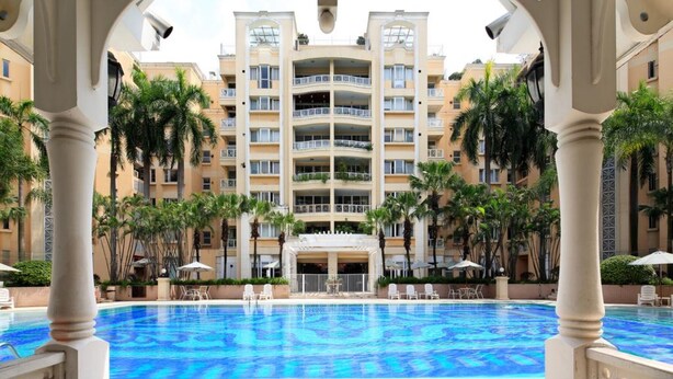 Ein Hotelpoolbereich mit klarem, blauem Pool, umgeben von Palmen und einem mehrstöckigen Gebäude im Hintergrund.