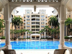 Une piscine d’hôtel à l’eau d’un bleu limpide, entourée de palmiers et avec un immeuble à plusieurs étages en arrière-plan.