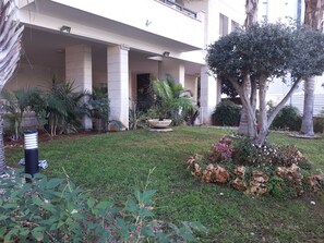 Un bâtiment doté d’un porche, de colonnes et d’un jardin où poussent différentes espèces de plantes et un arbre.