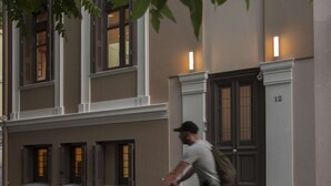 A man riding a bicycle past a building with multiple windows and a door marked with the number 12. - Tripodon Plaka in the shade of Acropolis by GHH (Athens)
