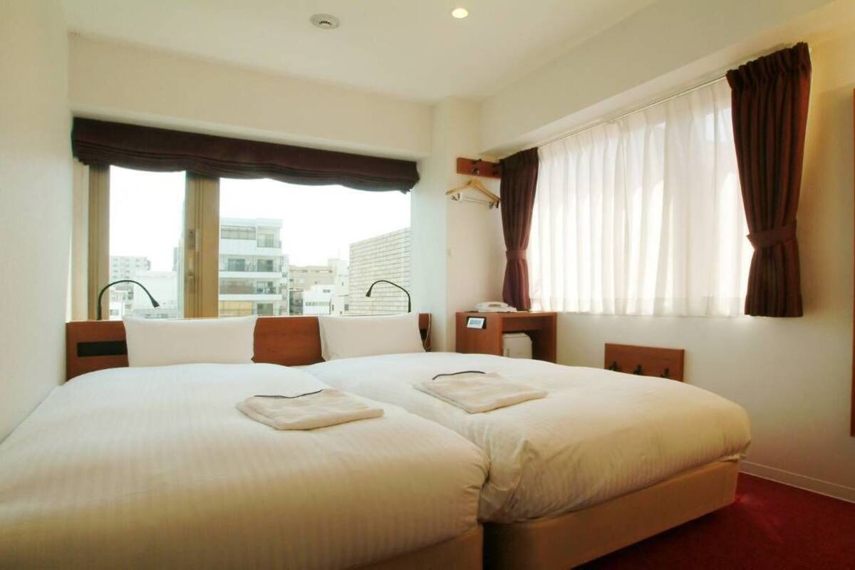 Ein Hotelzimmer mit zwei Betten, einem großen Fenster mit Vorhängen, einem Spiegel und Blick auf Gebäude.